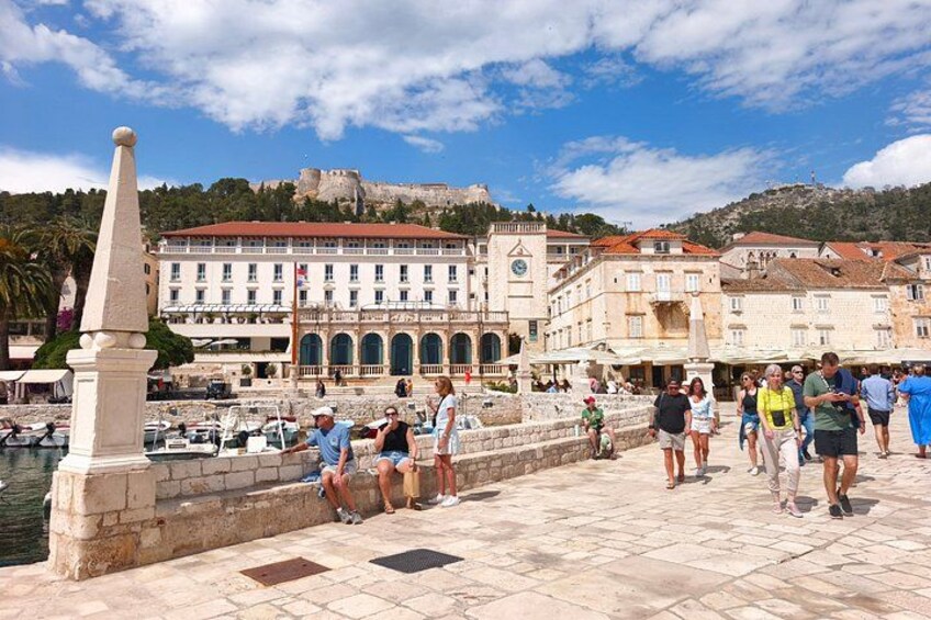 Un antico lungomare con persone che passeggiano, un edificio importante con una torre dell'orologio e una statua. 4