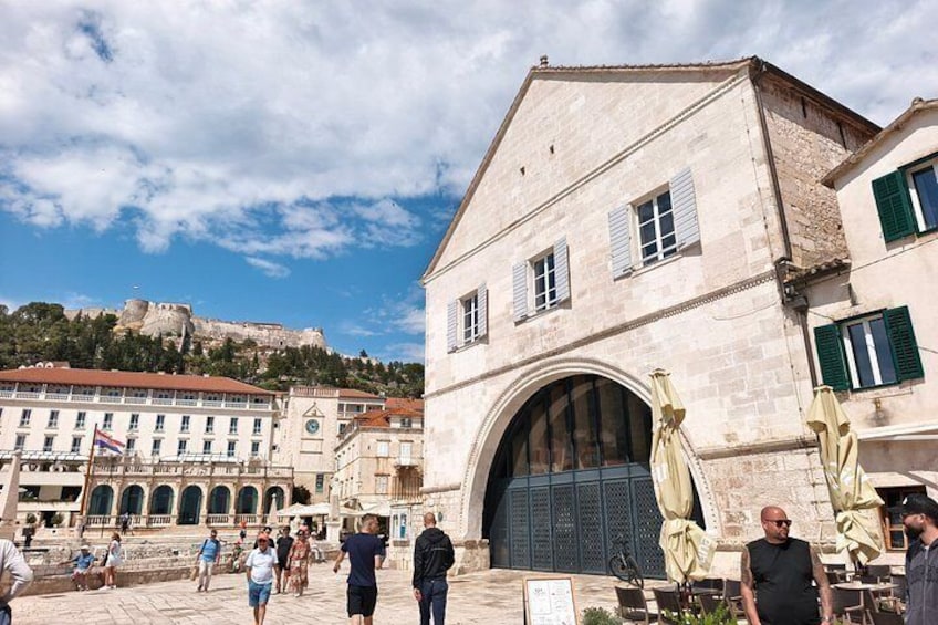 A historic building with a large arched entrance, a statue, and a view of a hilltop fortress in the background. 1