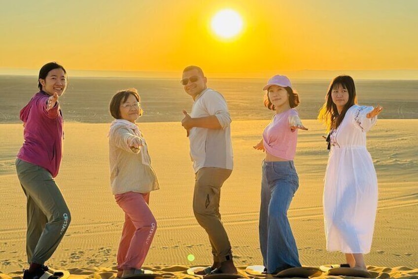 A group of people standing on a beach with the sun setting in the background. 1
