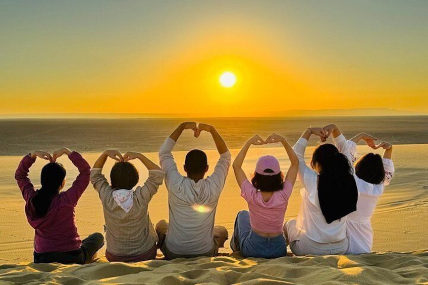Un grupo de personas sentadas en la playa al atardecer, formando un corazón con las manos. 5