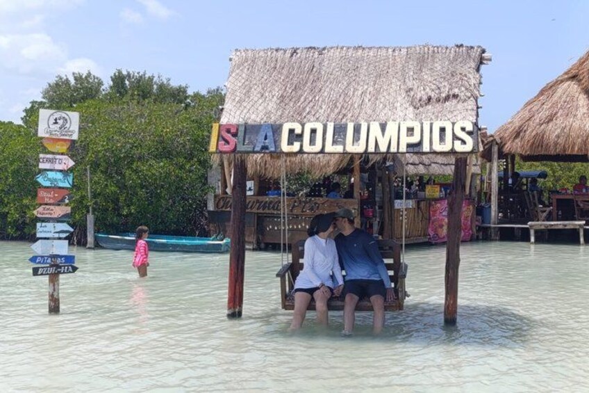 Una estructura de madera con techo de paja, identificada con el letrero 'ISLA COLUMPIOS', un poste de indicaciones con varios nombres de destinos y un bote pequeño al fondo. 4