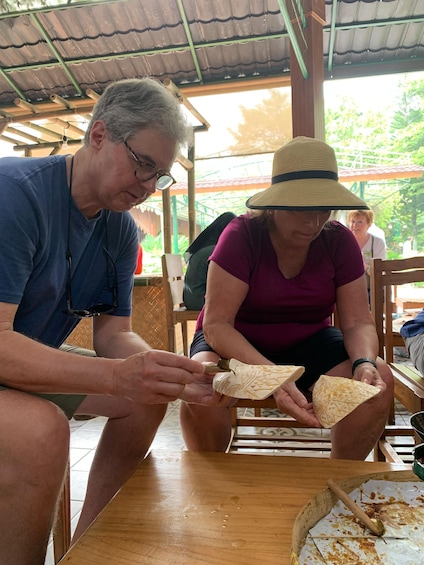 Two people seated at a wooden table under a thatched roof, engaged in a food preparation activity. 2