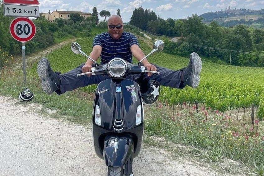 A person sitting on a scooter with their legs spread wide, a speed limit sign, and a rural landscape with vineyards. 4