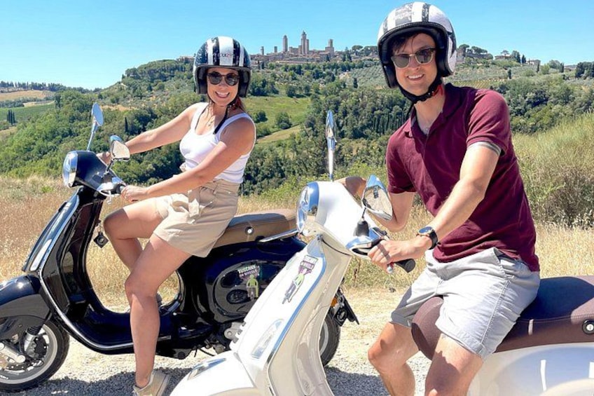 Two people on scooters in a grassy field with a hilly landscape in the background. 1