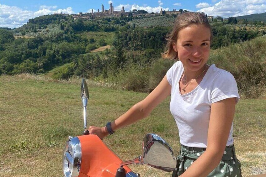 Una persona conduce un scooter rojo en un campo cubierto de hierba, con un paisaje montañoso y un castillo al fondo. 3