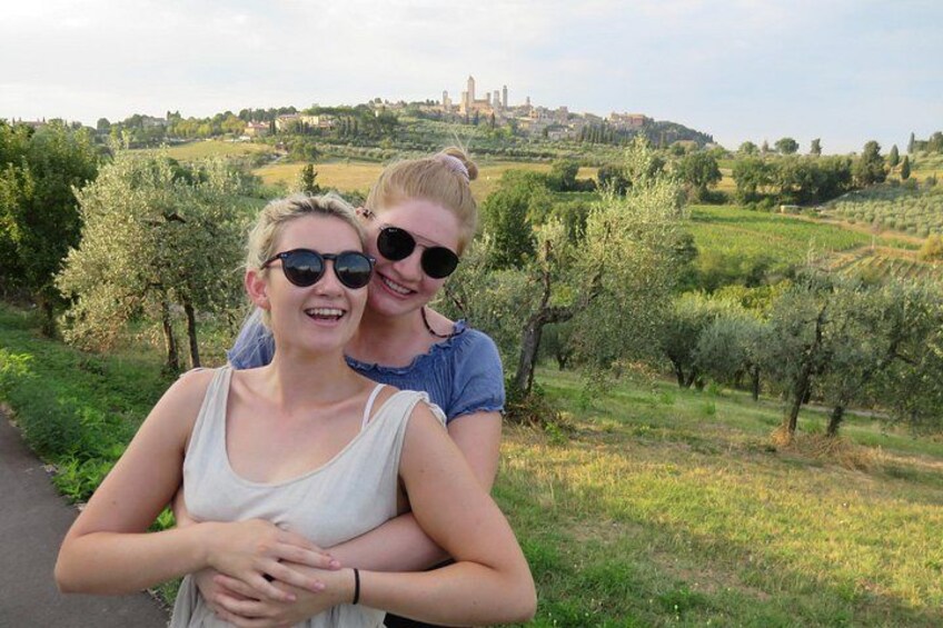 Two people standing in a vineyard with rows of olive trees and a distant view of a hilltop town. 2