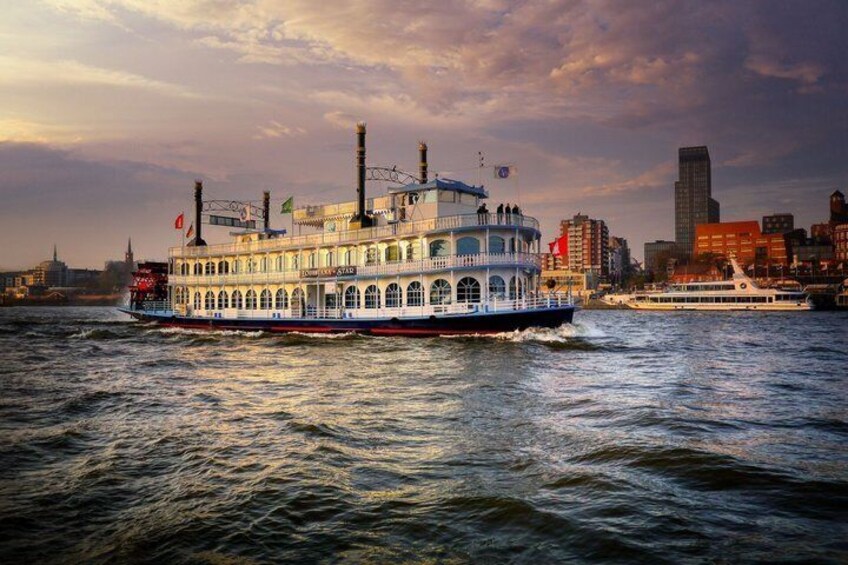 A riverboat with multiple decks and smokestacks, traveling on a river with a city skyline in the background. 1