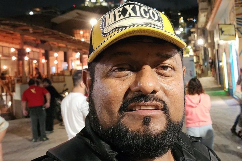 A person wearing a cap with 'MEXICO' written on it, standing on a street with shops and people in the background. 1