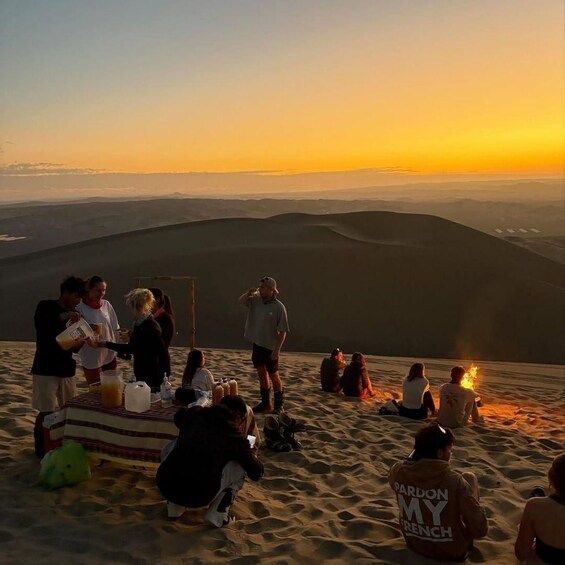 A group of people sitting and standing on a sand dune at sunset. 5