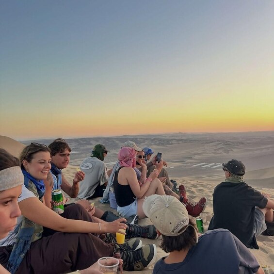 A group of people sitting on a sandy beach, some holding drinks, with a desert landscape in the background. 3