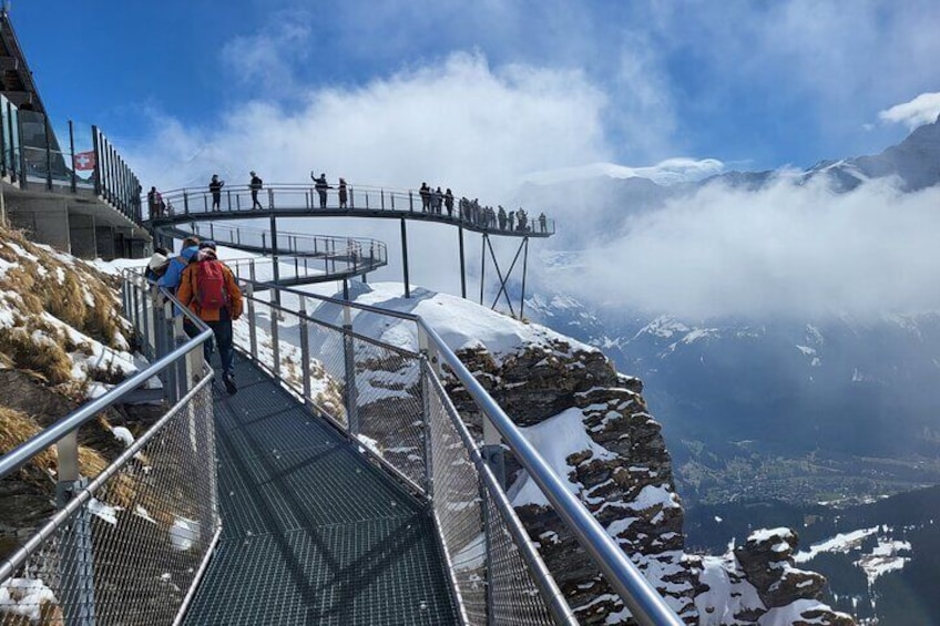 Eine Metallgangway mit Spaziergängern, mit Blick auf ein verschneites Bergmassiv und ein Tal. 1