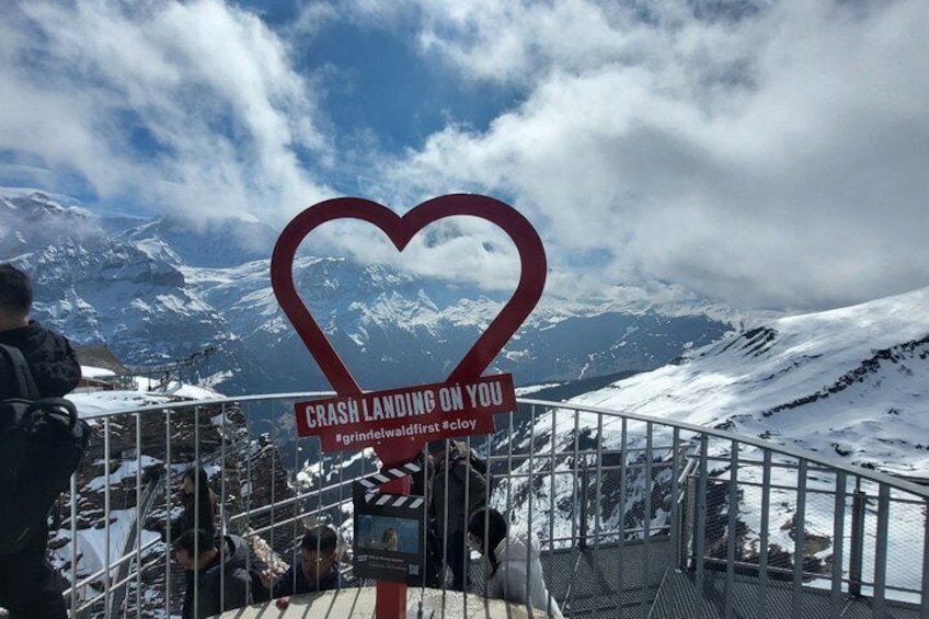 Eine Berglandschaft mit einem herzförmigen Schild, auf dem 'CRASH LANDING ON YOU' steht, und dem Hashtag '#grindelwaldfirst'. 3