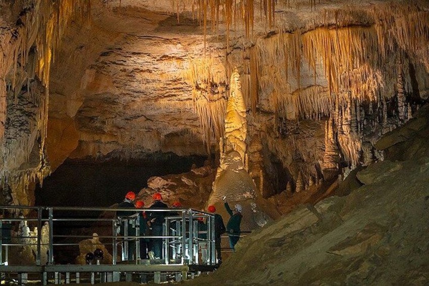 A cave with stalactites and stalagmites, a group of people wearing helmets and harnesses, and a metal walkway. 1