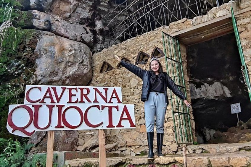 A person standing in front of a sign that reads 'CAVERNA QUIOCTA' with a stone wall and a green gate in the background. 2
