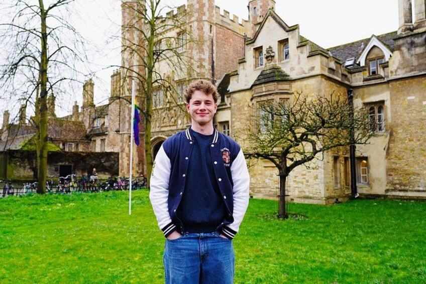 A person standing in front of a historic building with stone architecture, a flagpole, and a tree in a grassy area. 4