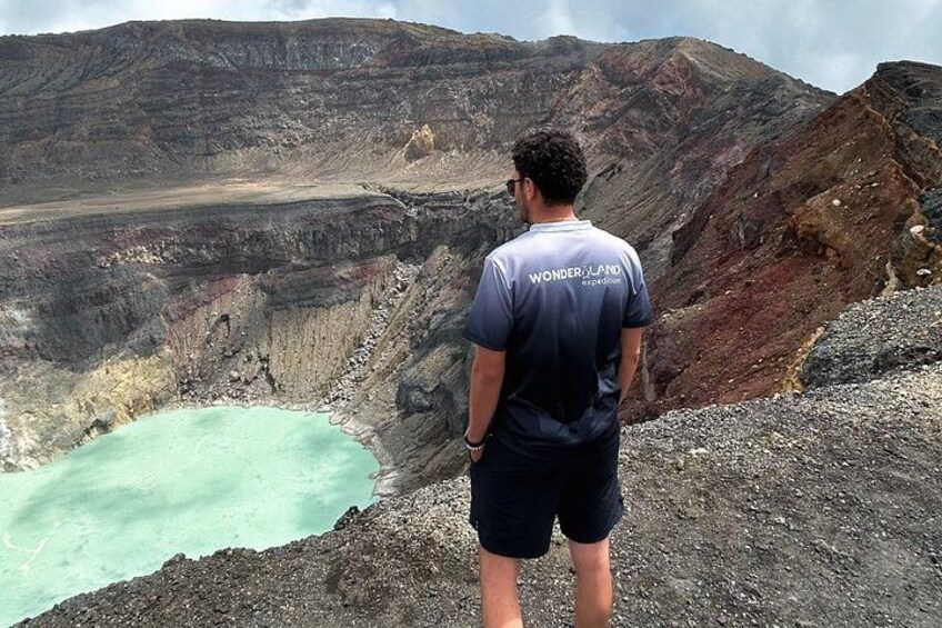 A person standing on a rocky outcrop overlooking a crater with a turquoise lake. 1