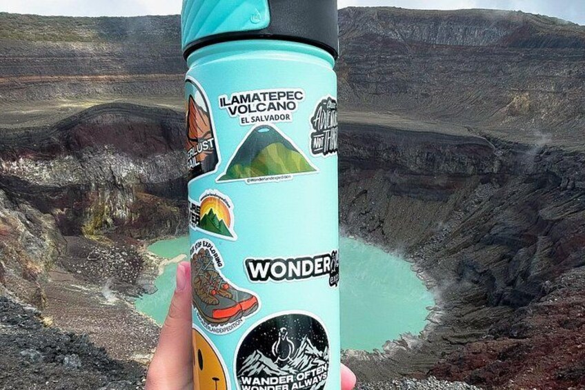 A hand holding a water bottle with a volcano-themed design, set against a backdrop of a volcanic crater. 3