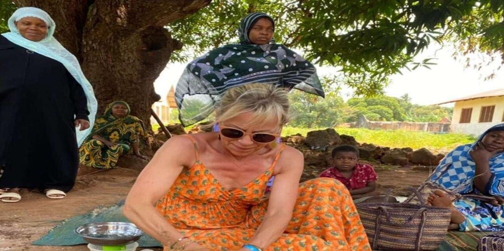A group of people sitting outdoors under a tree, with a woman in an orange dress in the foreground. 3