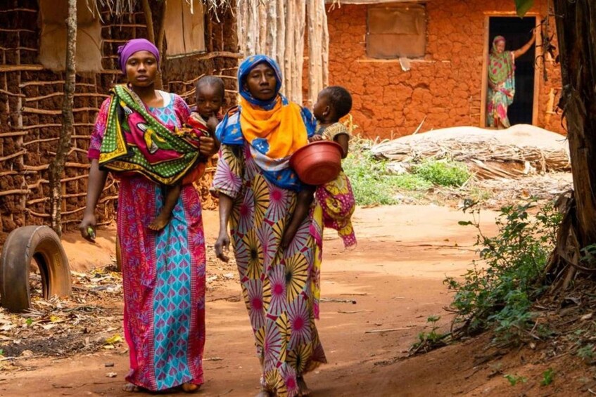 A rural village scene with women and children walking along a dirt path, traditional houses, and a tire in the background. 2