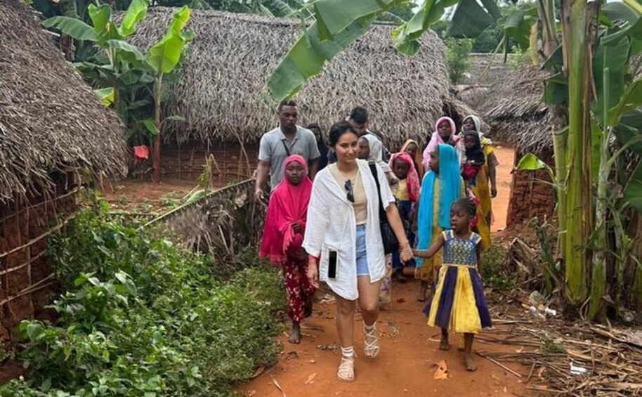 A group of people walking through a rural area with thatched-roof huts and banana plants. 5