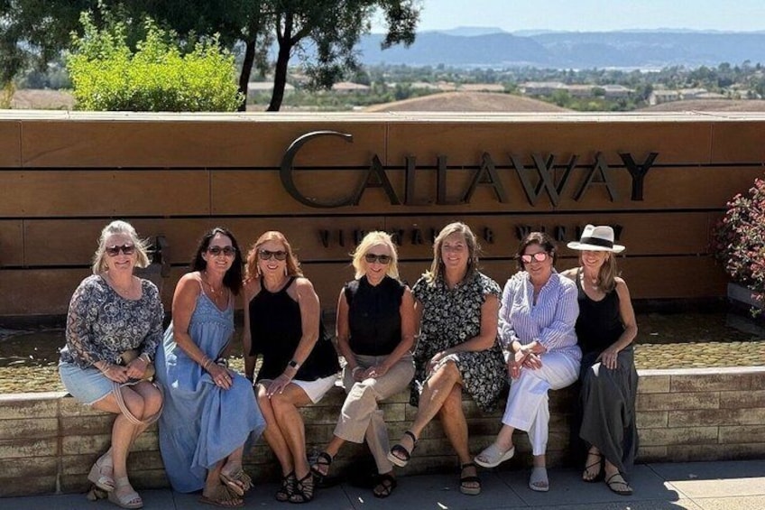 A group of people sitting on a bench in front of a sign that reads 'Caliway.' 1