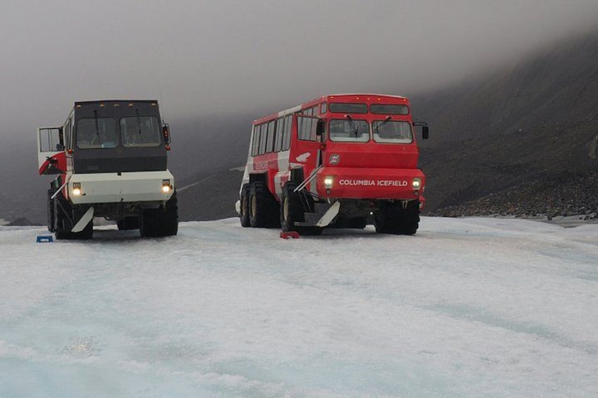 To røde køretøjer med 'COLUMBIA ICEFIELD' skrevet på, parkeret på en sneklædt overflade med et bjerglandskab i baggrunden. 1