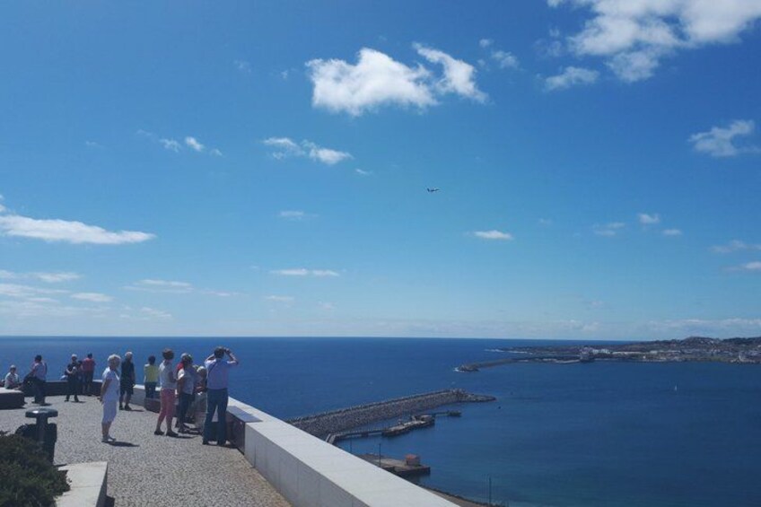 Een kustlandschap met mensen die op een promenade staan en uitkijken over de zee, een vliegtuig in de lucht en een steiger die het water in reikt. 4