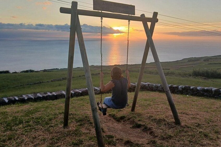 Une personne assise sur une balançoire en bois, d’où l’on peut admirer un coucher de soleil sur une étendue d’eau. 3
