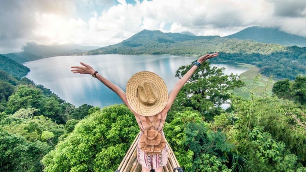 A person with arms raised, wearing a straw hat, overlooking a lake surrounded by lush greenery and mountains. 1
