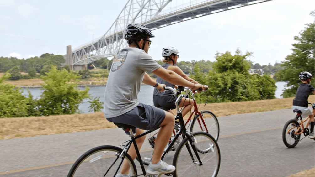Three cyclists on a paved path with a bridge and greenery in the background. 1