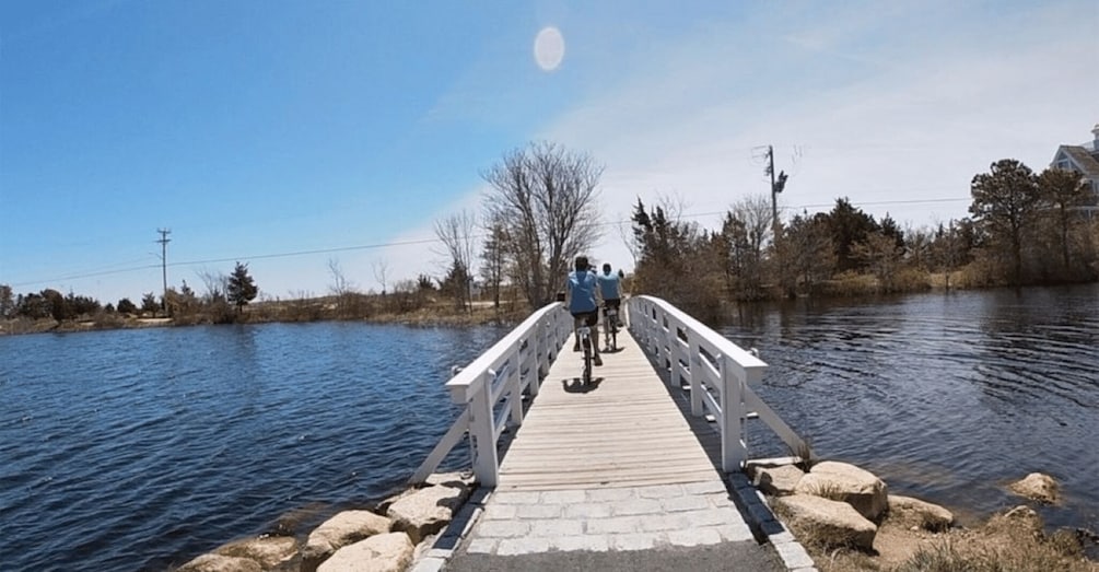A wooden bridge over a calm river with two people cycling, surrounded by trees and a clear sky. 3