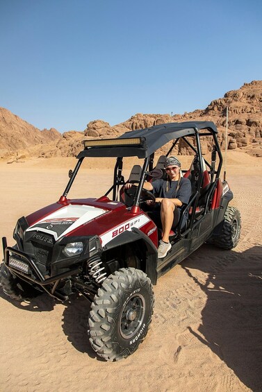 A red and white utility vehicle with a person seated inside, parked on a sandy terrain with rocky hills in the background. 1