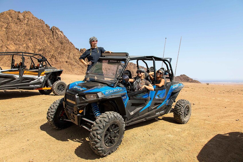 A blue Polaris 1000 HD off-road vehicle with passengers, parked on a desert with rocky terrain in the background. 5