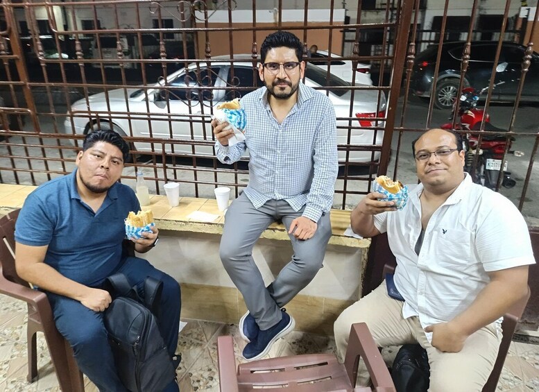 Three men sitting on chairs, each holding a wrapped snack, with a car and motorcycles in the background. 2