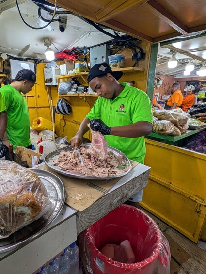 A person in a green shirt preparing food at a counter with a large metal tray of meat, a red trash bin, and various food items in the background. 3
