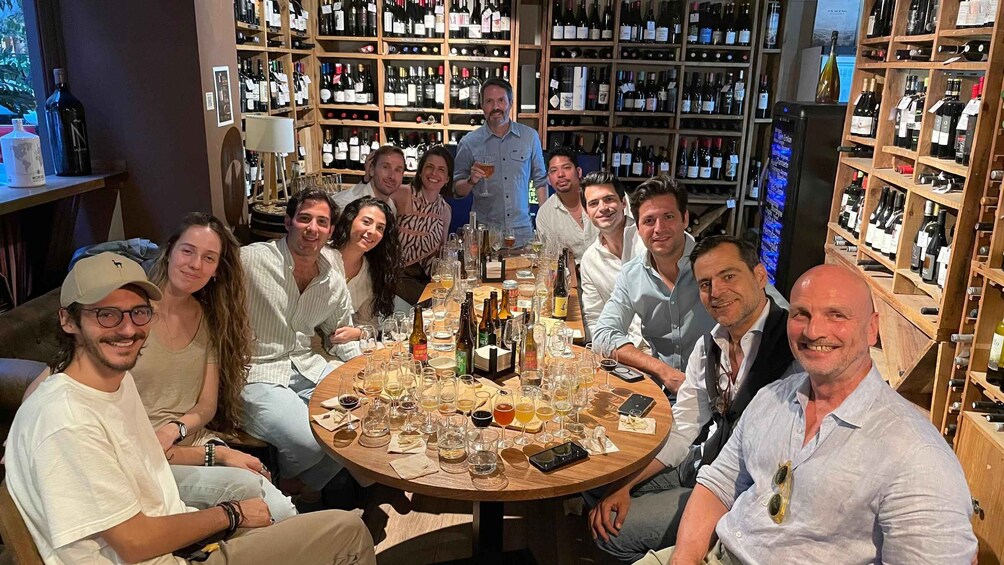 A group of people seated around a table with various bottles and glasses, in a room with shelves of wine bottles in the background. 1