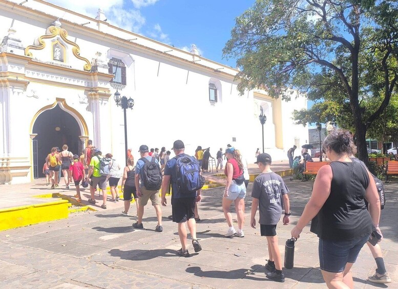 A group of people walking on a paved street in front of a white building with ornate architectural details. 2
