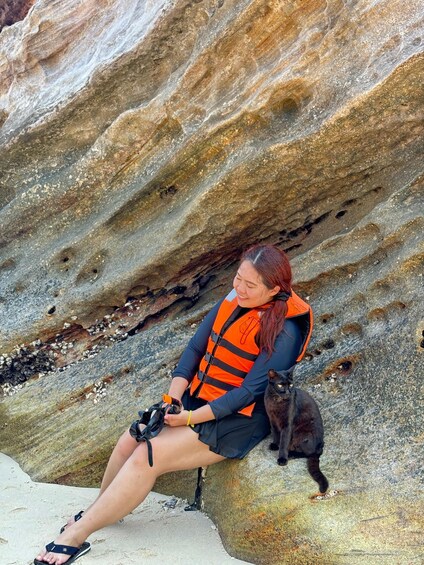 A person sitting on a rock with a black cat, wearing an orange life jacket. 2