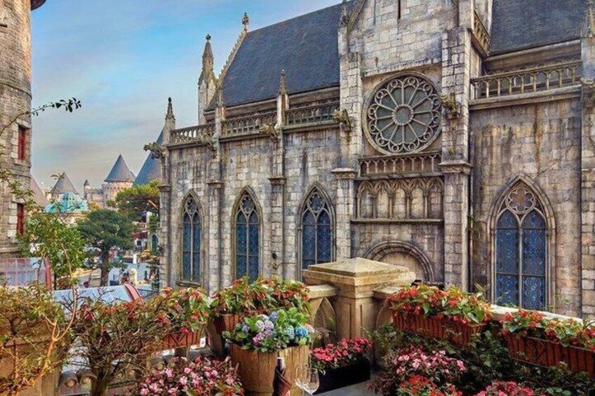 A historic stone building with arched windows and a large circular window, surrounded by a garden with flowering plants. 5