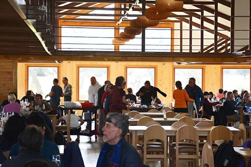 A crowded indoor dining area with wooden tables and chairs, large windows, and hanging light fixtures. 4