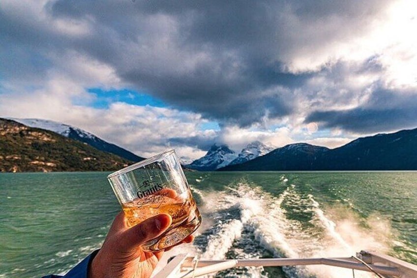 En hand som håller i ett glas med dryck, en båt på vattnet och berg i bakgrunden. 2