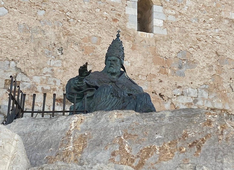 Une statue représentant une figure drapée, tenant une croix, placée devant un mur de pierre percé d’une petite fenêtre. 2
