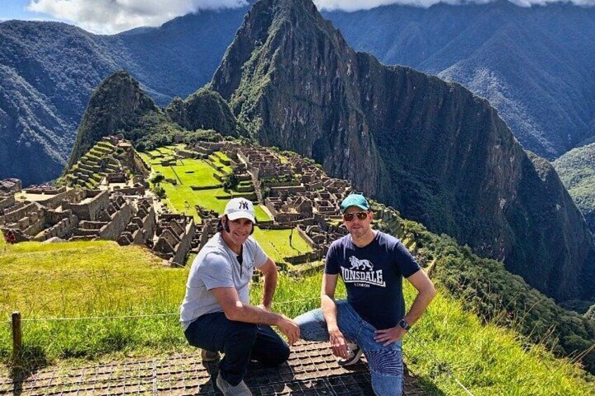 Two people standing in front of Machu Picchu with the ruins and surrounding mountains in the background. 1