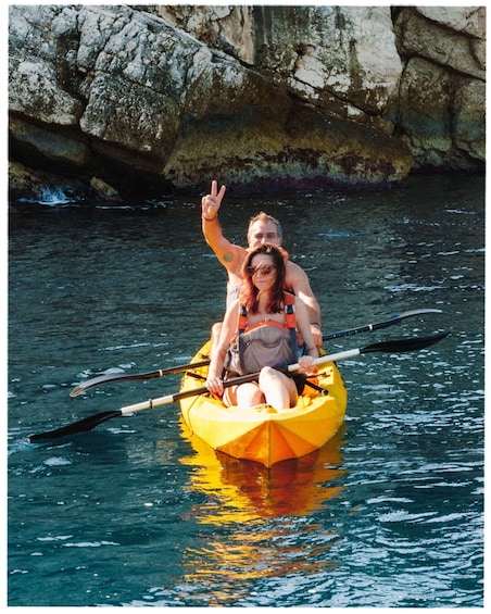 Two people in a yellow kayak on calm water with a rocky shoreline in the background. 2