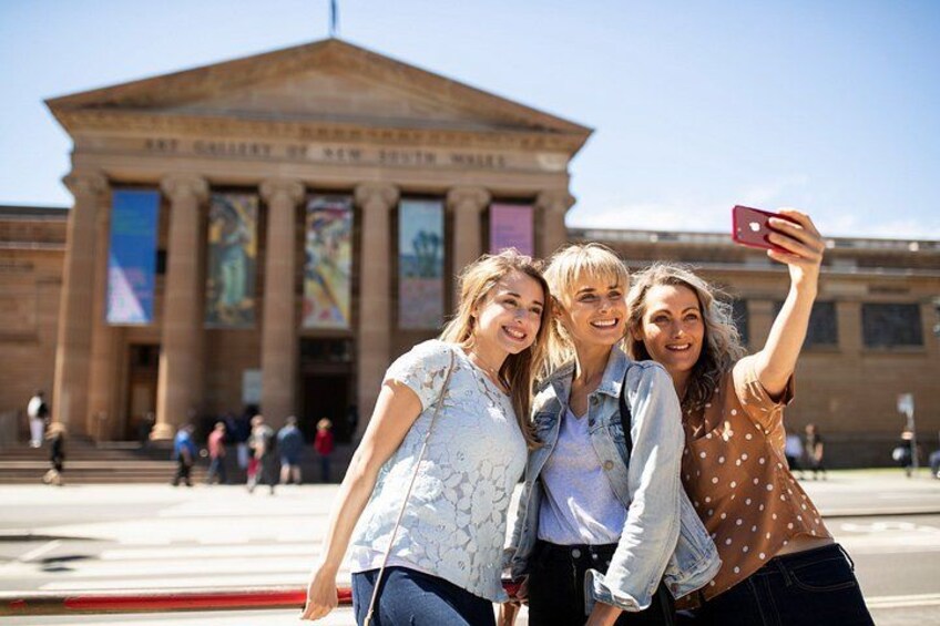 Tres personas tomándose un selfie frente a un edificio clásico con columnas. 1