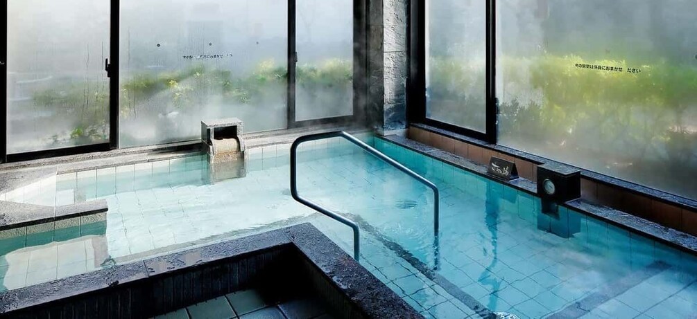 An indoor swimming pool with clear glass walls, a tiled floor, and a small seating area. 1
