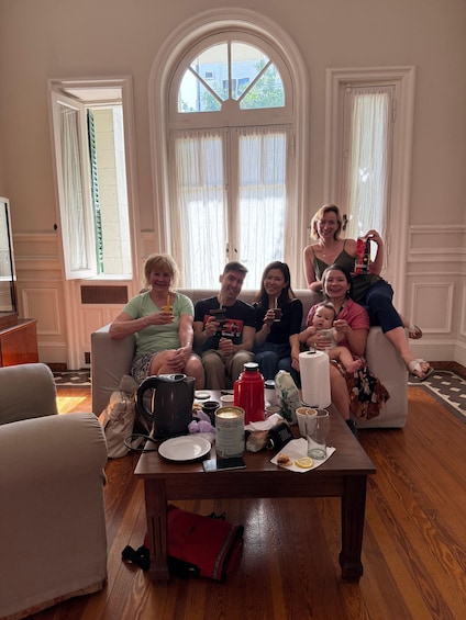 A group of people sitting on a couch in a room with a large window, a table with various items, and a red backpack on the floor. 3