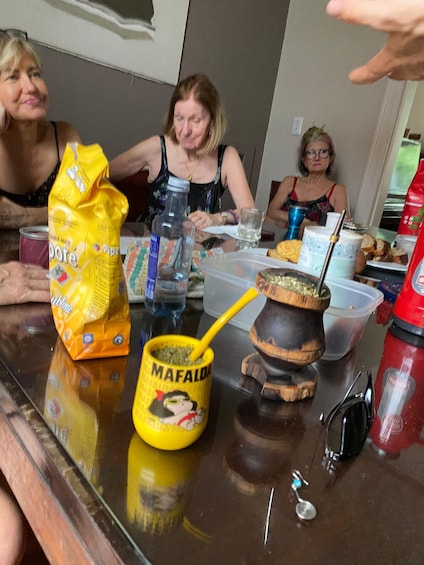 A group of people sitting at a table with a yellow mug labeled 'MAFALDA,' a plastic bag, a bottle, and a red fire extinguisher. 2