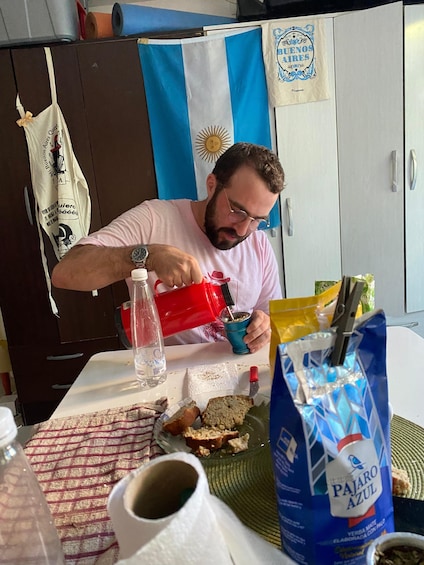 A person pouring a beverage into a cup, with a flag, a bag of coffee, and bread on the table. 1