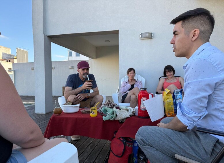 A group of people sitting at a table on a rooftop terrace with a red tablecloth, enjoying a meal. 5